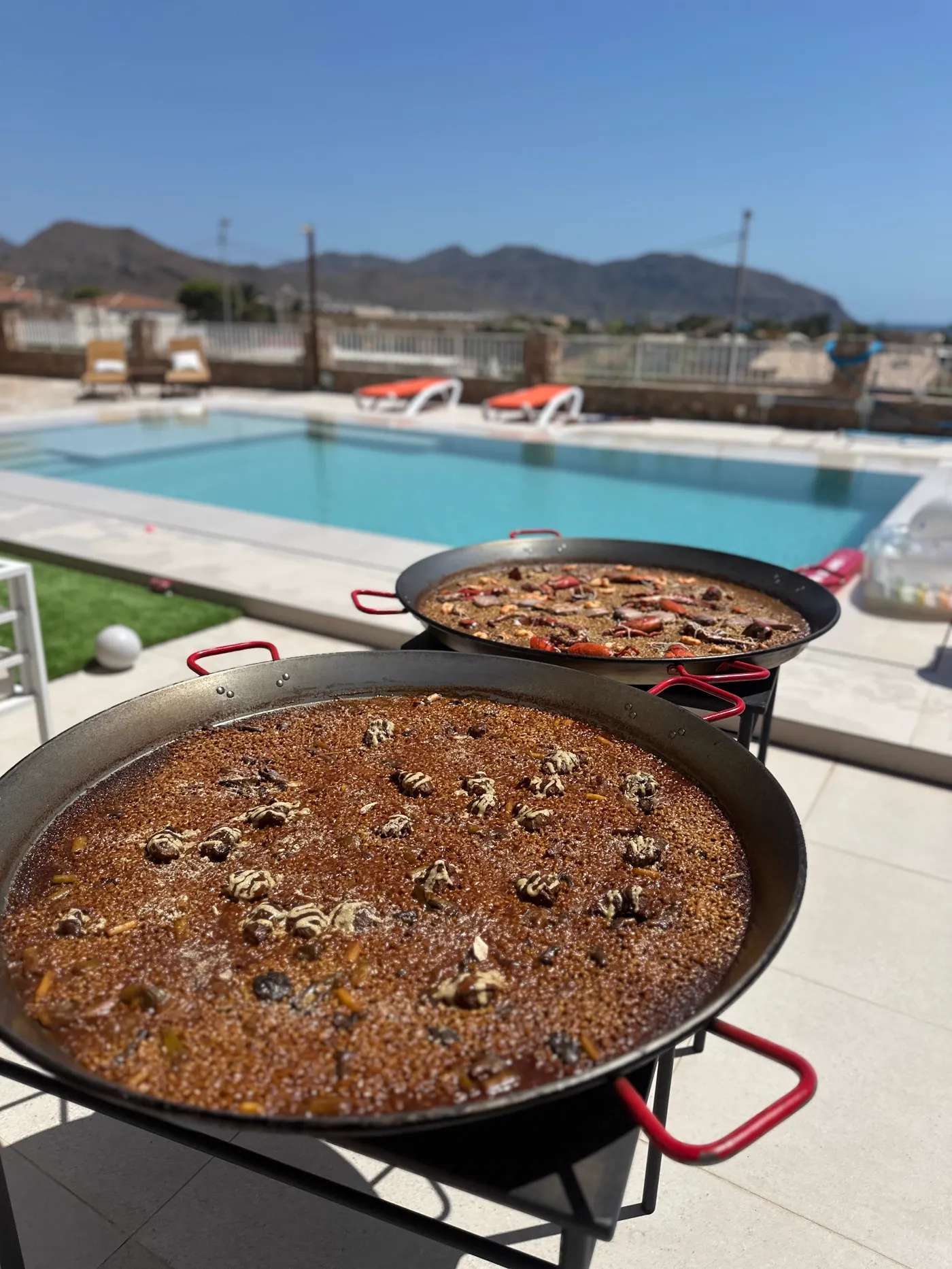 Paellas al sol junto a una piscina con el monte detrás